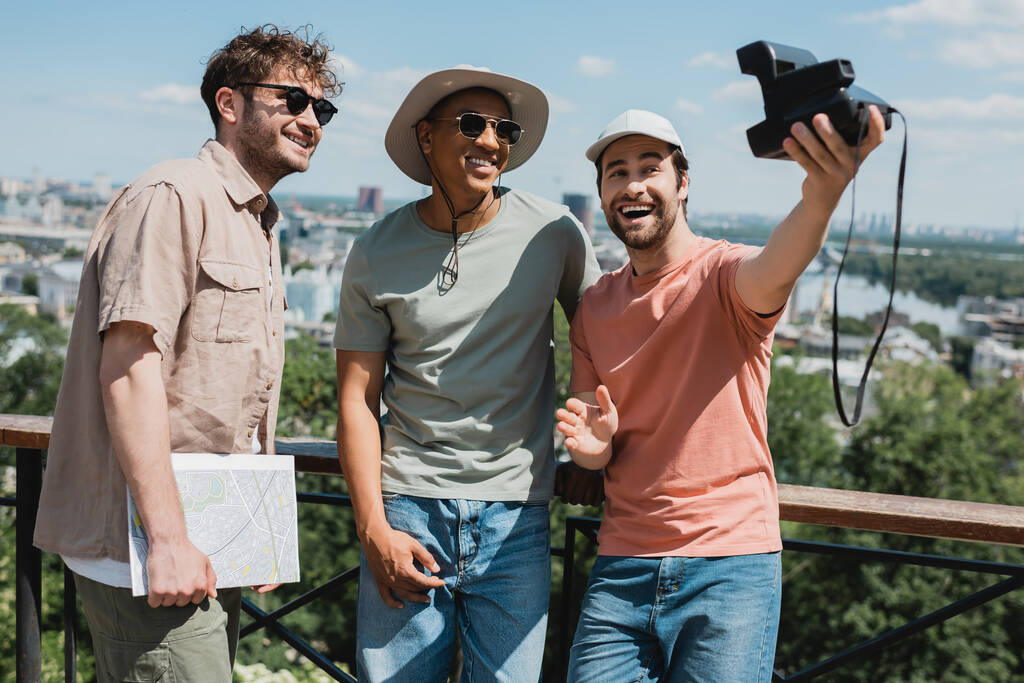 spensierato uomo barbuto con fotocamera vintage scattare selfie con amico africano americano e guida turistica vicino alla recinzione nel parco della città - Foto, immagini