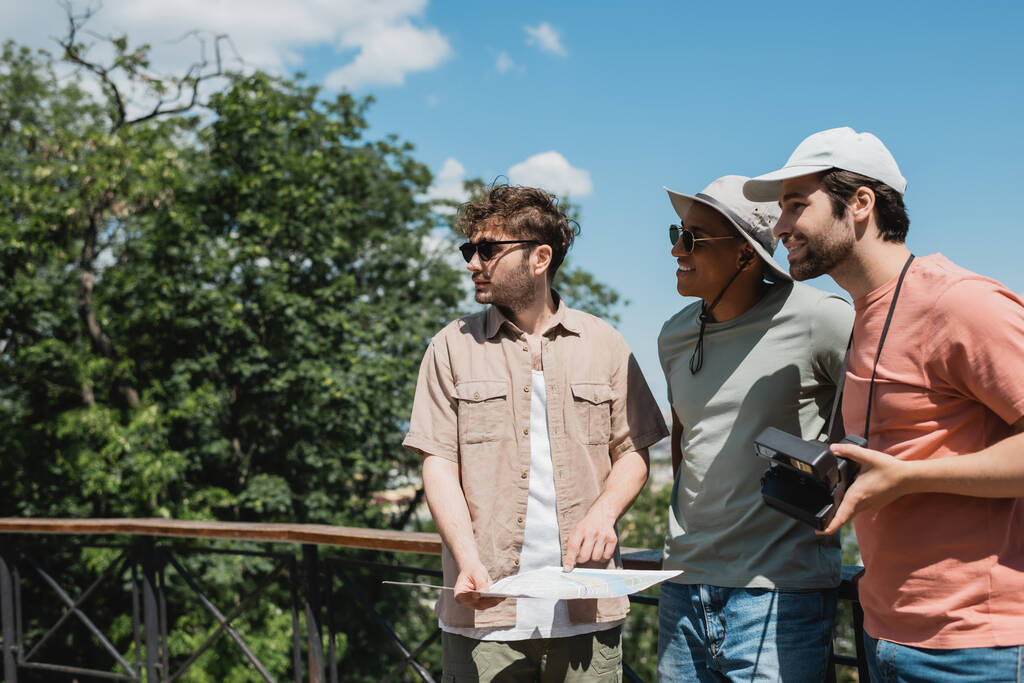 счастливые мультиэтнические путешественники в солнечных шляпах глядя в сторону рядом с экскурсоводом с картой в городском парке - Фото, изображение