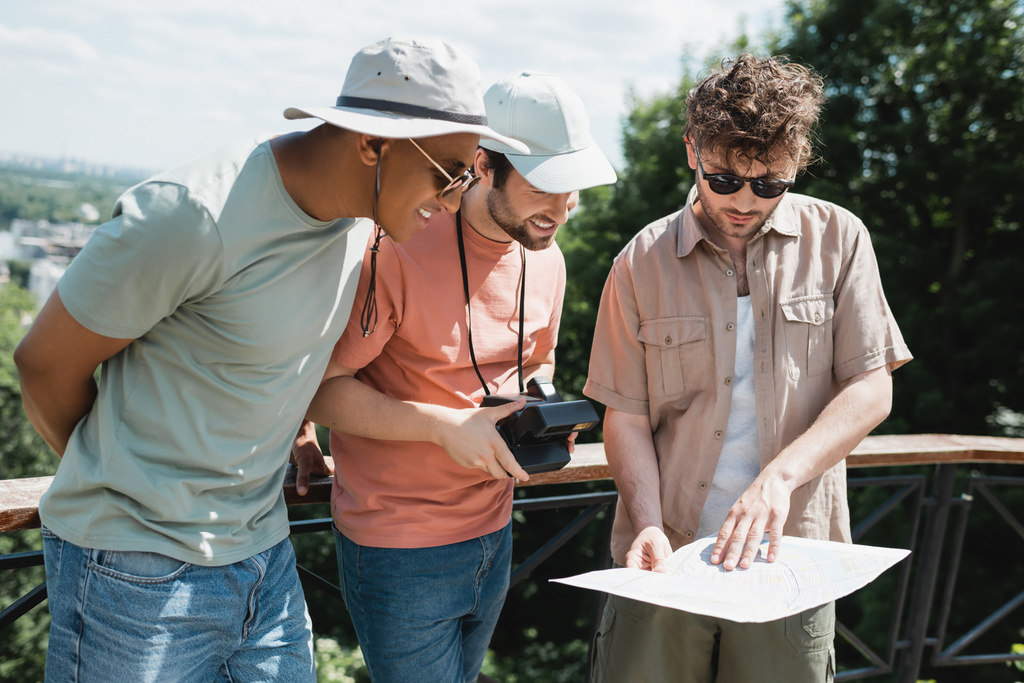 Güneş gözlüklü genç tur rehberi şehir parkındaki gezi sırasında güneş şapkalı ırklar arası turistlerin haritasını gösteriyor. - Fotoğraf, Görsel