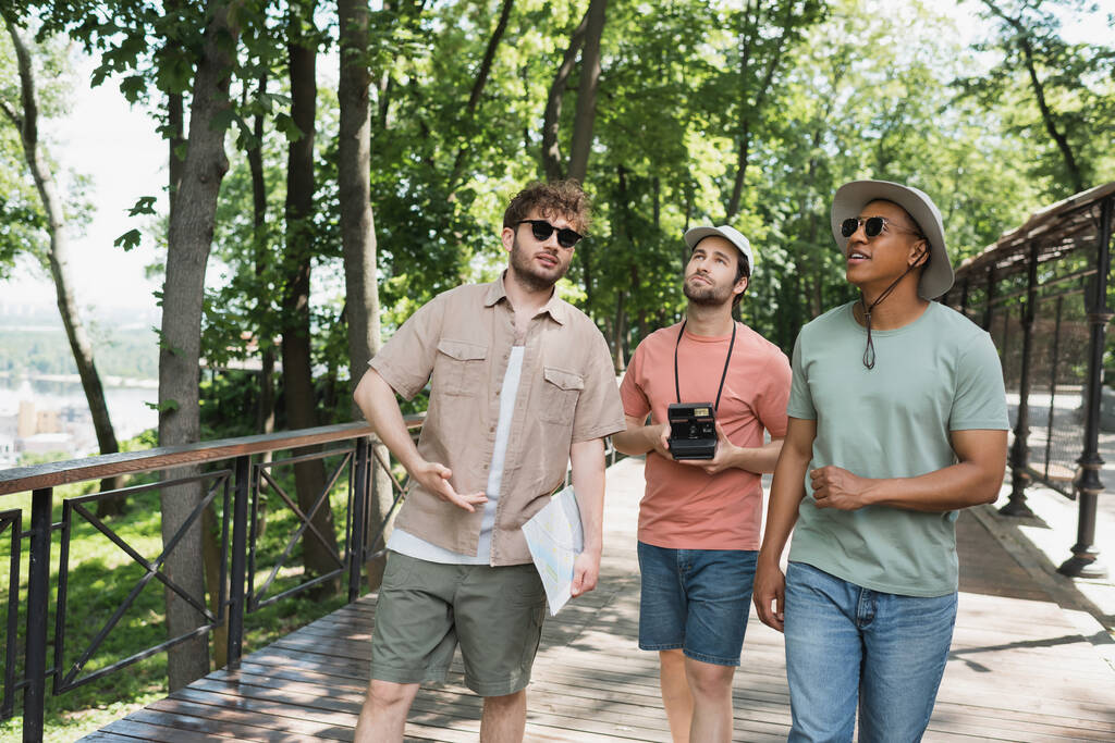 multicultural travelers in sun hats looking away during summer walk with tour guide in urban park - Photo, Image