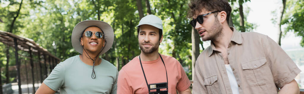 молодой гид в солнечных очках разговаривает с многонациональными туристами в солнечных шляпах в зеленом парке, баннер - Фото, изображение
