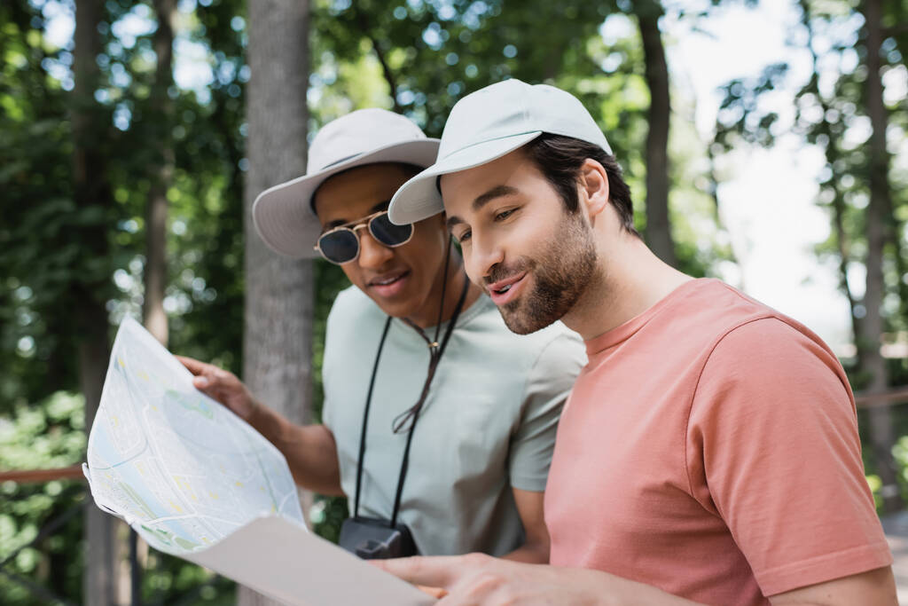 веселі багатоетнічні туристи в сонячних капелюхах дивляться на карту подорожей в розмитому парку
 - Фото, зображення