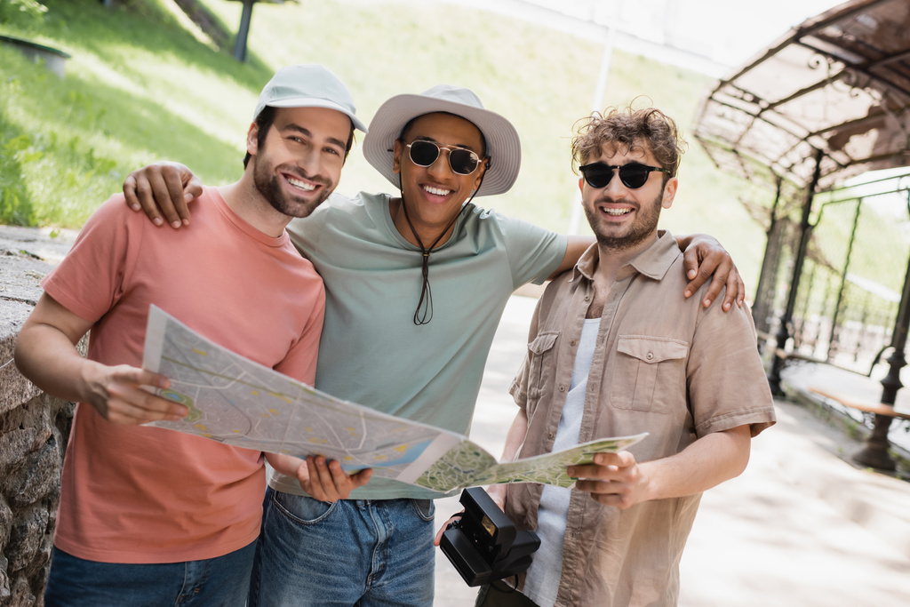 despreocupado hombre afroamericano en sombrero de sol y gafas de sol abrazando amigos en la calle urbana - Foto, imagen