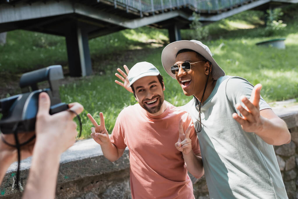 размытый человек с винтажной камерой фотографирует беззаботных межрасовых друзей, позирующих в солнечных шляпах и жестикулирующих в городском парке - Фото, изображение