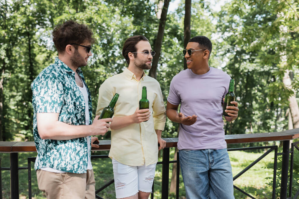 amigos multiétnicos felices y elegantes en gafas de sol con cerveza fresca y hablando cerca de valla en el parque de la ciudad - Foto, Imagen
