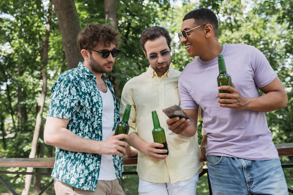 uomini alla moda che tengono la birra e guardando smartphone in mano di felice amico afroamericano nel parco della città - Foto, immagini