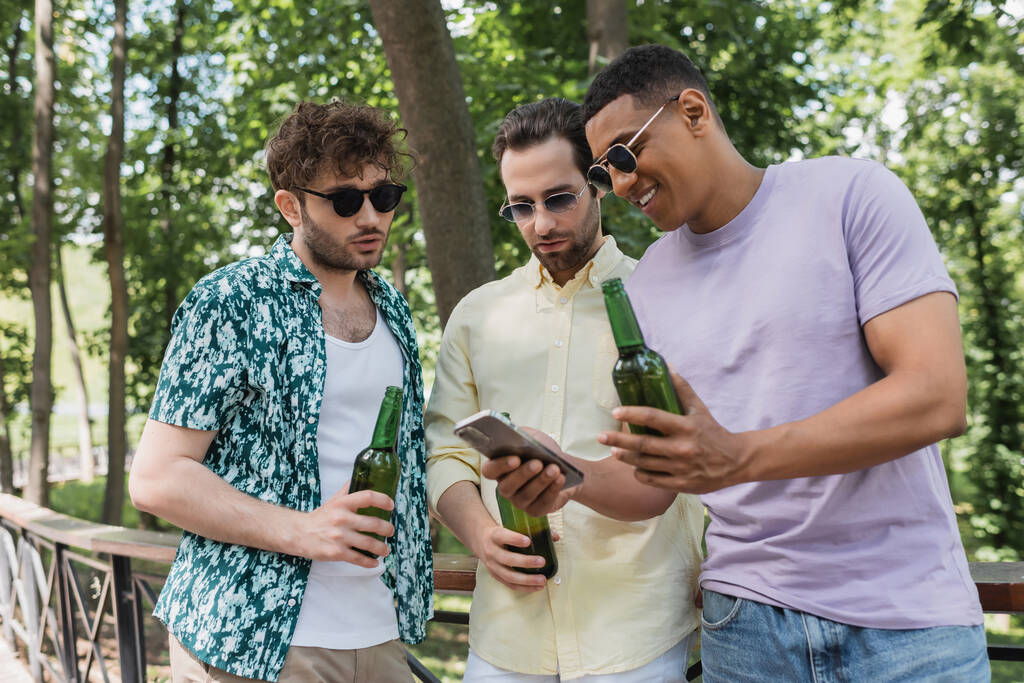 wesoły Afroamerykanin w okularach przeciwsłonecznych pokazujący telefon komórkowy modnym przyjaciołom trzymającym piwo w parku miejskim - Zdjęcie, obraz