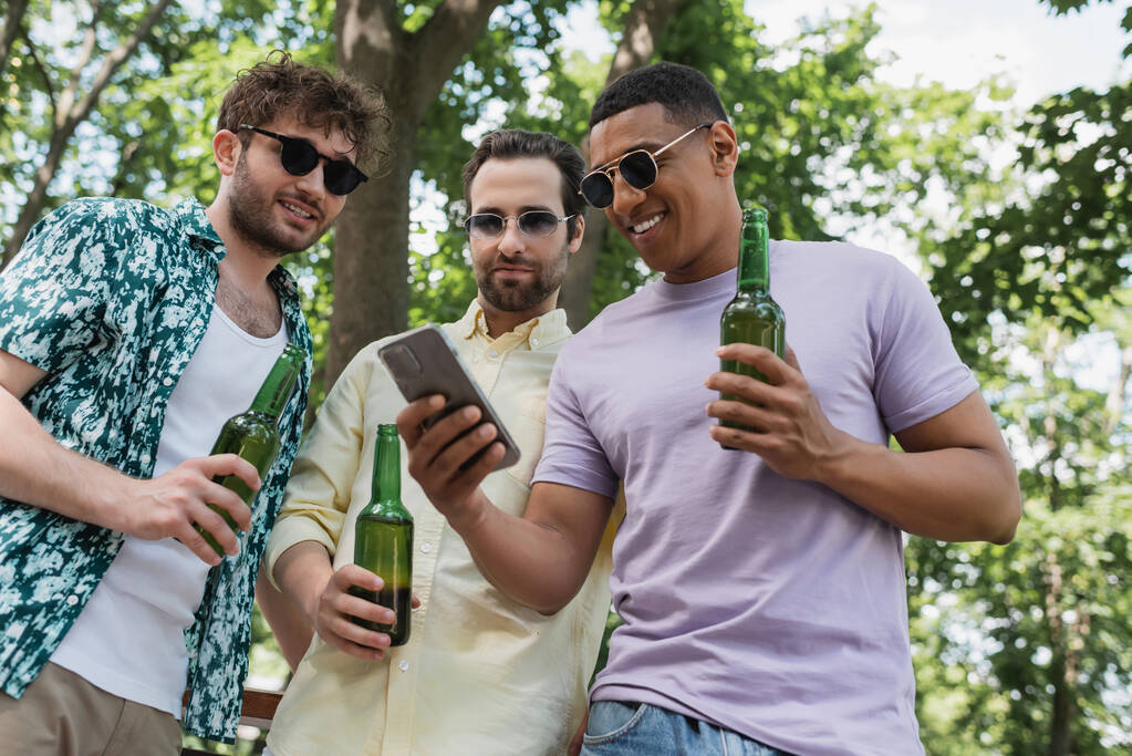 радісний афроамериканський чоловік тримає мобільний телефон поруч зі стильними друзями з пляшками пива в парку
 - Фото, зображення