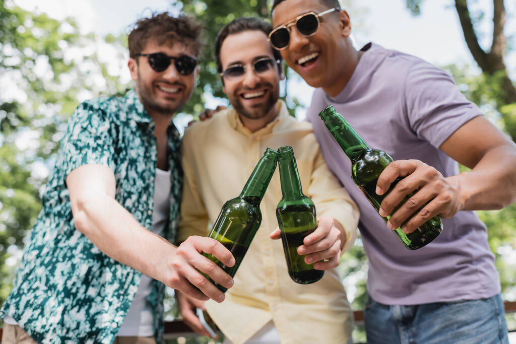 Unbekümmert und stilvoll multiethnische Männer mit Sonnenbrille klimpern Bierflaschen im Park - Foto, Bild