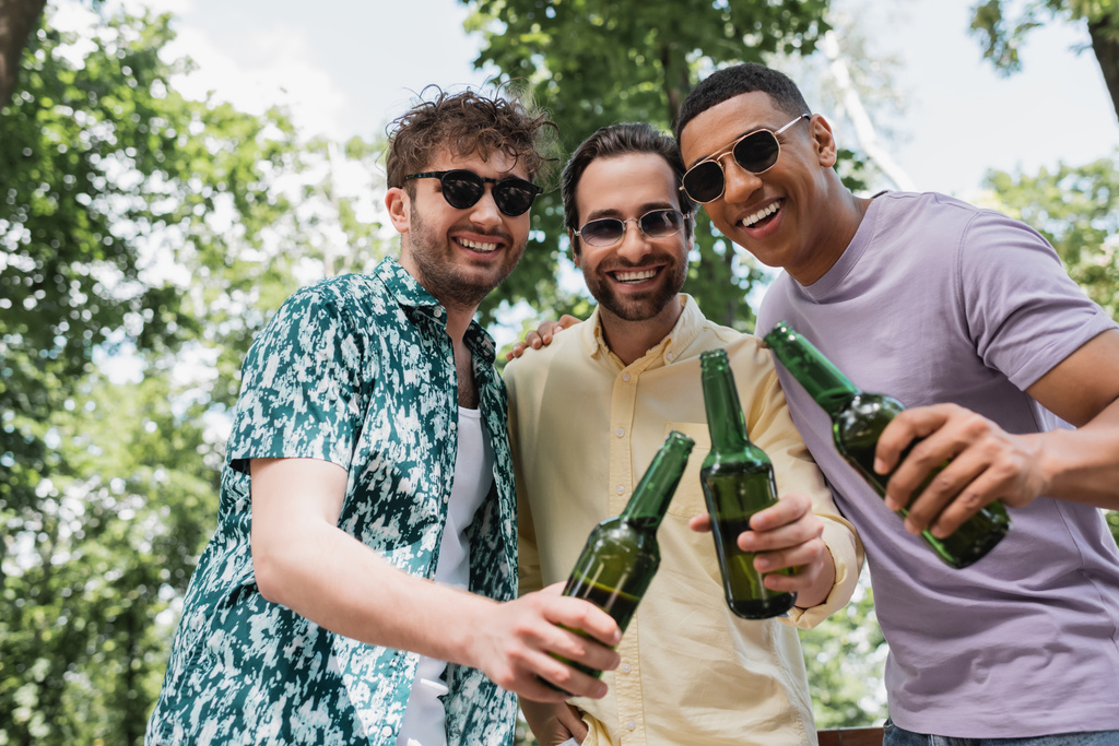 iloinen monikulttuurinen ystäviä trendikkäitä kesävaatteita ja aurinkolasit paahtaminen tuoretta olutta vihreässä puistossa - Valokuva, kuva