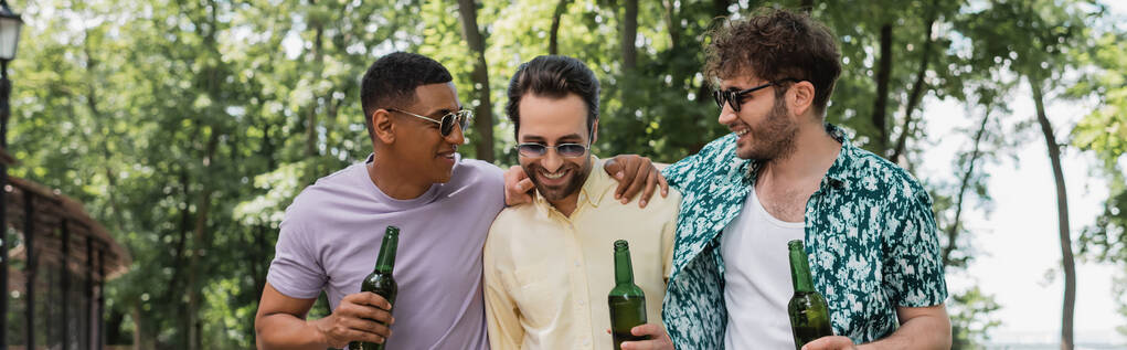 amigos multiétnicos alegres y de moda sosteniendo la cerveza y abrazando mientras pasa tiempo en el parque de verano, pancarta - Foto, imagen