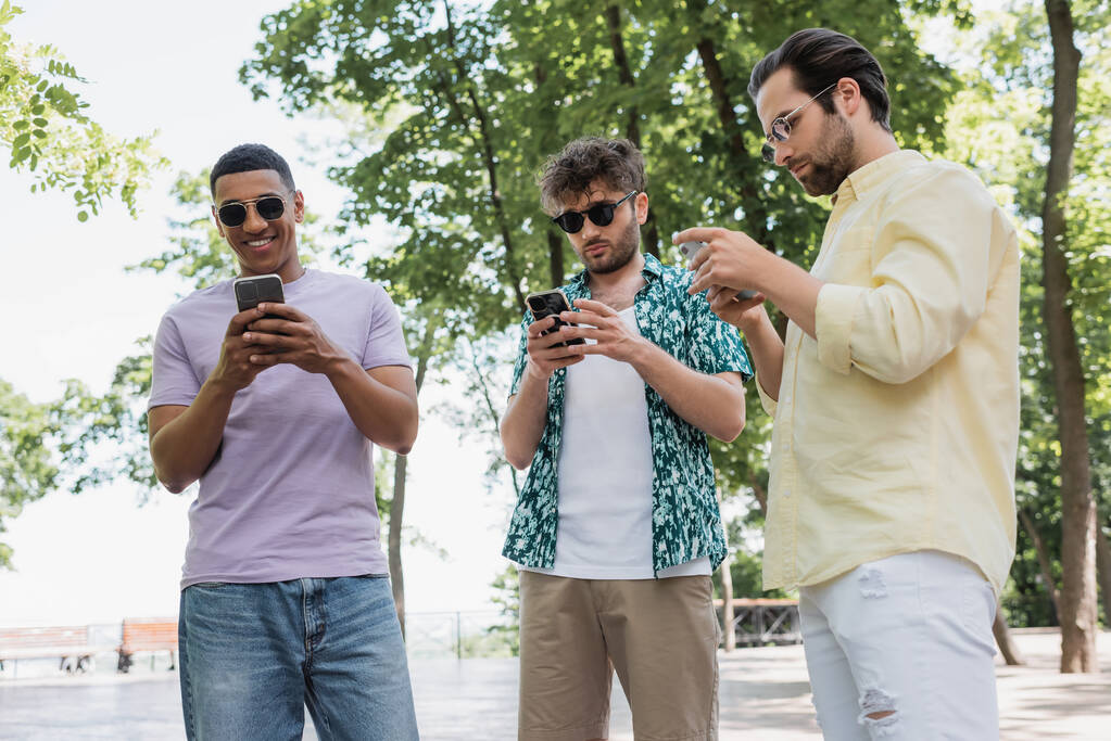 uomini multiculturali in elegante abito estivo e occhiali da sole utilizzando smartphone nel parco cittadino - Foto, immagini