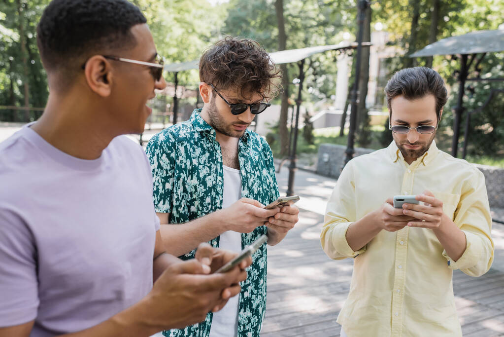 Afrikanisch-amerikanischer Mann schaut trendigen Freunden beim Handy-Chatten im Sommerpark zu - Foto, Bild