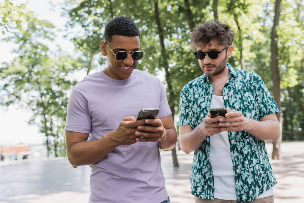 Χαμογελώντας διαφυλετικούς φίλους σε γυαλιά ηλίου χρησιμοποιώντας smartphones στο πάρκο  - Φωτογραφία, εικόνα
