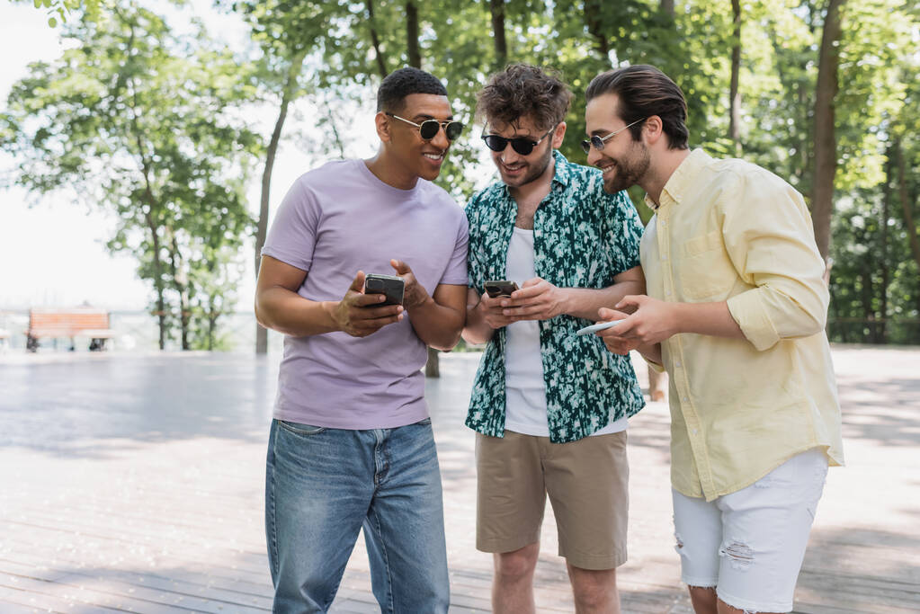 Allegro uomo afroamericano che usa lo smartphone e parla con gli amici in occhiali da sole nel parco  - Foto, immagini