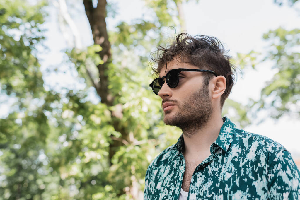 Mladý muž v slunečních brýlích a košili stojící v rozmazaném letním parku  - Fotografie, Obrázek