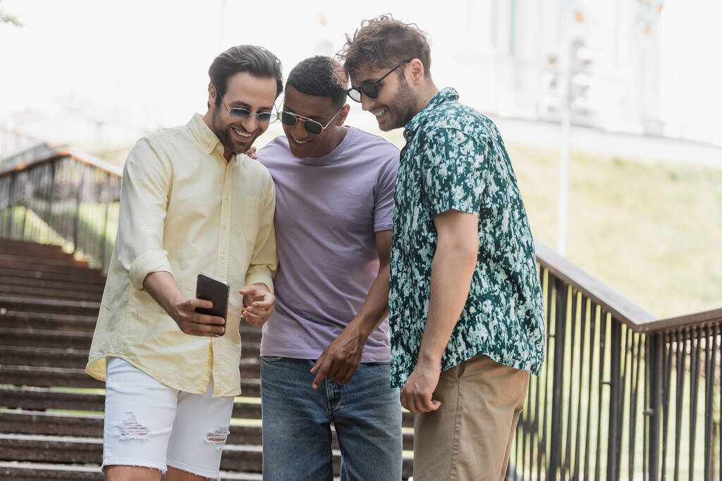 Joven mostrando teléfono celular a amigos multiétnicos en gafas de sol en las escaleras en el parque  - Foto, Imagen
