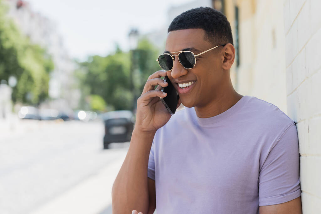 Homem americano africano alegre em óculos de sol falando no smartphone na rua urbana turva  - Foto, Imagem