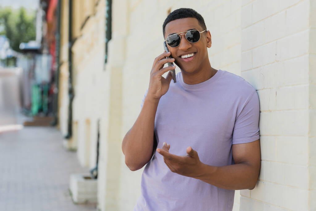 Sonriente hombre afroamericano en gafas de sol hablando por teléfono móvil en la calle urbana borrosa  - Foto, imagen