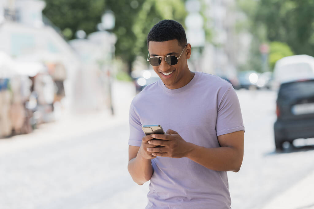 Hombre afroamericano despreocupado en gafas de sol usando teléfono móvil en la calle urbana  - Foto, Imagen
