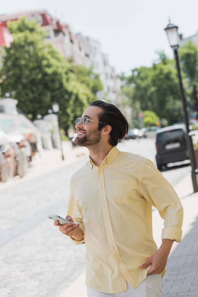 Positiver Mann mit Sonnenbrille benutzt Handy beim Gehen auf verschwommener Stadtstraße  - Foto, Bild