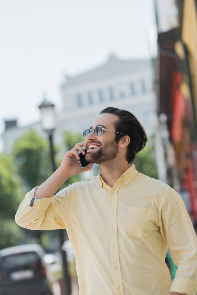Homme positif en lunettes de soleil et chemise parlant sur smartphone dans la rue urbaine  - Photo, image