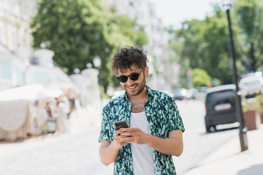 Nadšený muž v slunečních brýlích chůze a používání smartphone na městské ulici  - Fotografie, Obrázek