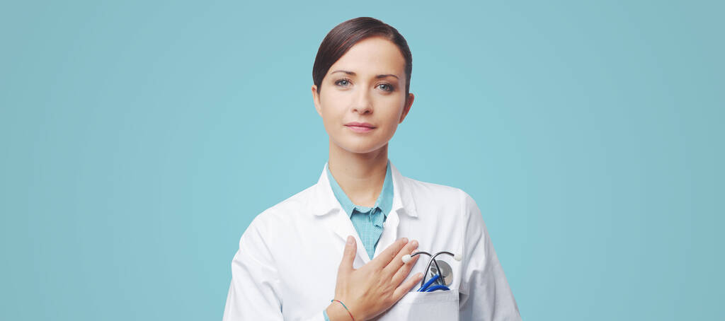 Attraente cardiologa femminile con stetoscopio e camice da laboratorio, che tocca il petto
. - Foto, immagini