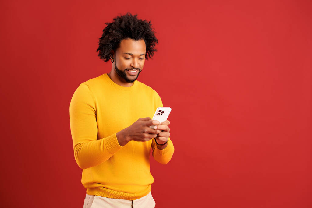 A vonzó szakállas férfi portréja, aki boldog arckifejezéssel mobiltelefont használ, okostelefon-függő, SMS-ezik a közösségi hálón. Fedett stúdió lövés elszigetelt piros háttér - Fotó, kép