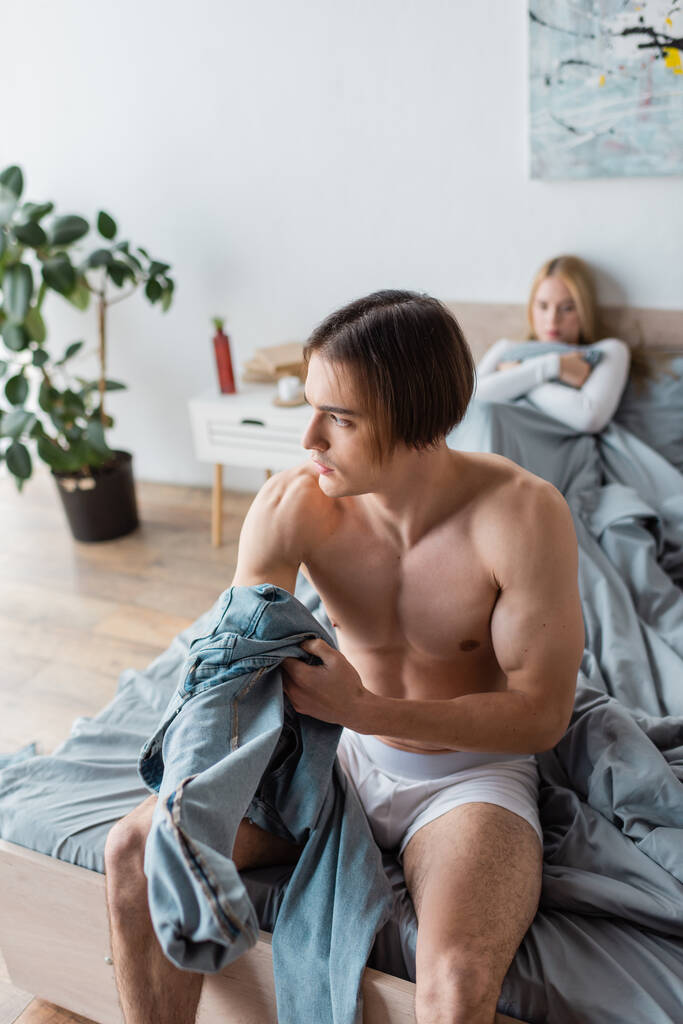 hombre sin camisa sosteniendo jeans mientras está sentado en la cama cerca de la mujer después de una noche de stand  - Foto, imagen