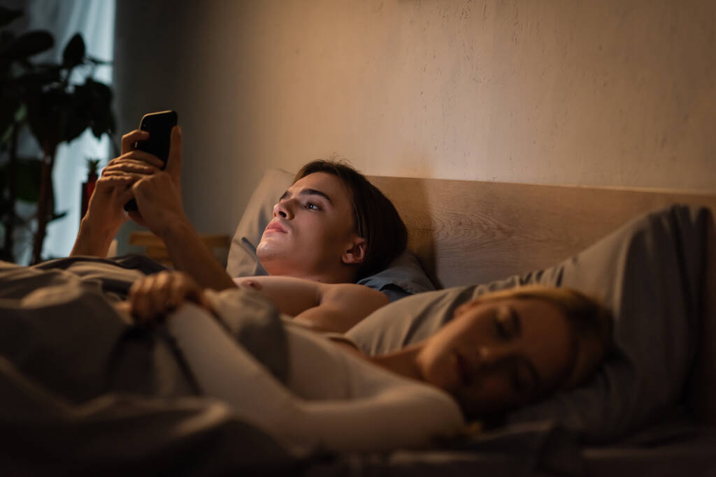 νεαρός άνδρας που χρησιμοποιεί το κινητό τηλέφωνο δίπλα σε ξανθιά φίλη κοιμάται στο κρεβάτι, εξαπάτηση έννοια  - Φωτογραφία, εικόνα