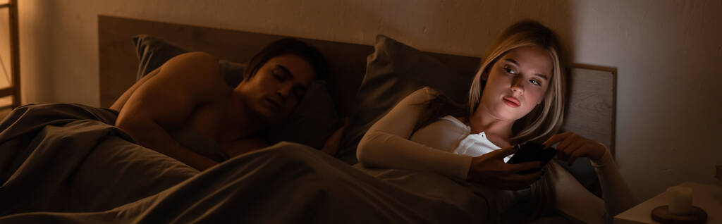 donna bionda messaggistica su smartphone accanto al fidanzato addormentato di notte, banner   - Foto, immagini
