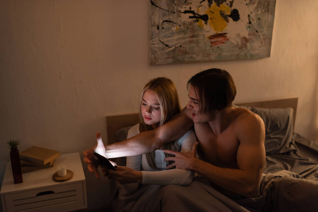 angry man screaming while arguing with blonde girlfriend holding smartphone in bedroom, cheating concept  - Photo, Image
