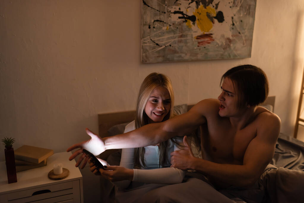 эмоциональный мужчина ссорится с обеспокоенной девушкой держа смартфон в спальне, измена концепции  - Фото, изображение