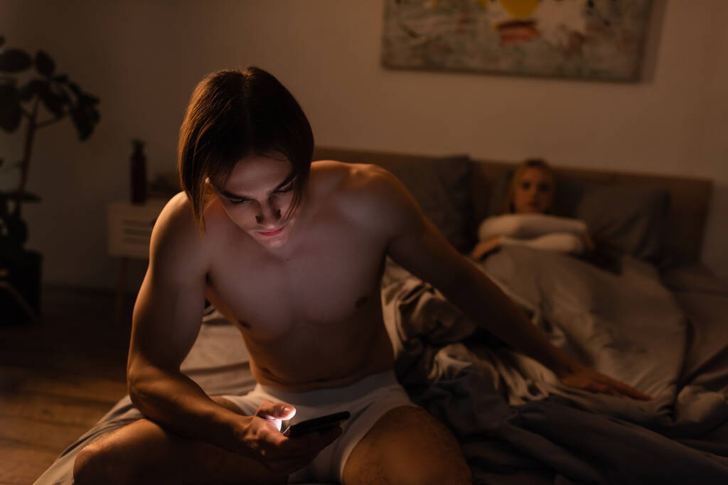 shirtless man using mobile phone while sitting on bed near girlfriend on blurred background, cheating concept  - Photo, Image