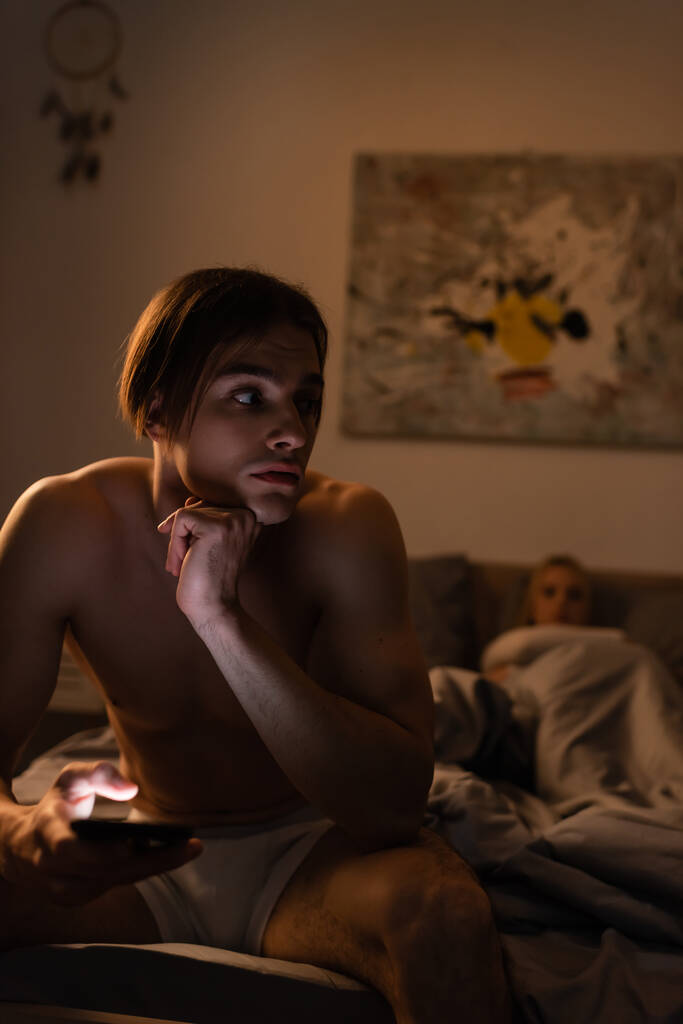 hombre sin camisa usando el teléfono móvil mientras está sentado junto a su novia en un fondo borroso, concepto de engaño  - Foto, Imagen