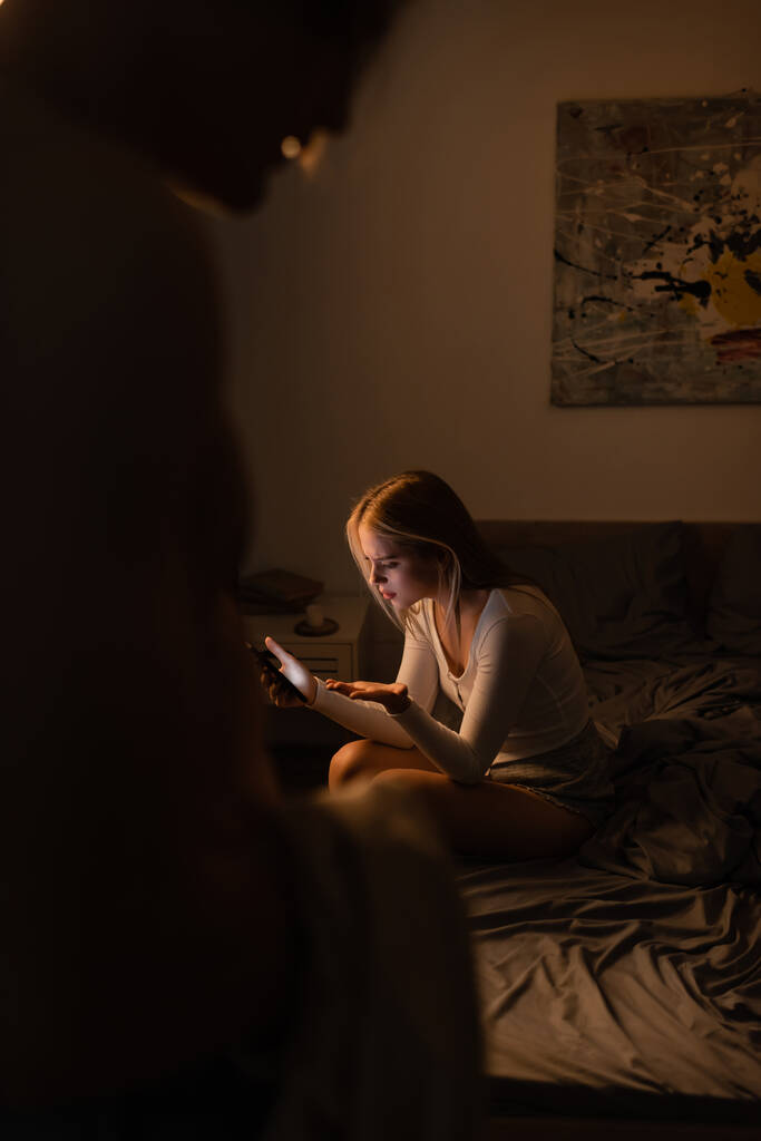 rozrušená žena čtení zpráv na smartphone v blízkosti rozmazané nevěrný přítel v noci  - Fotografie, Obrázek