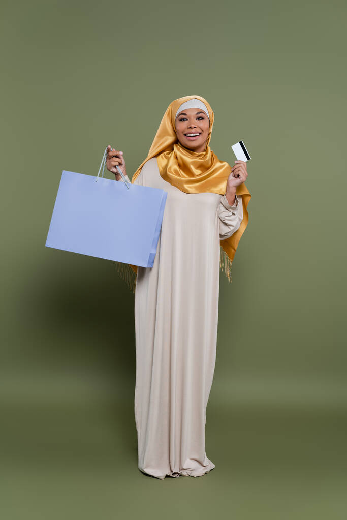 Mulher multirracial alegre no hijab segurando cartão de crédito e saco de compras em fundo verde - Foto, Imagem