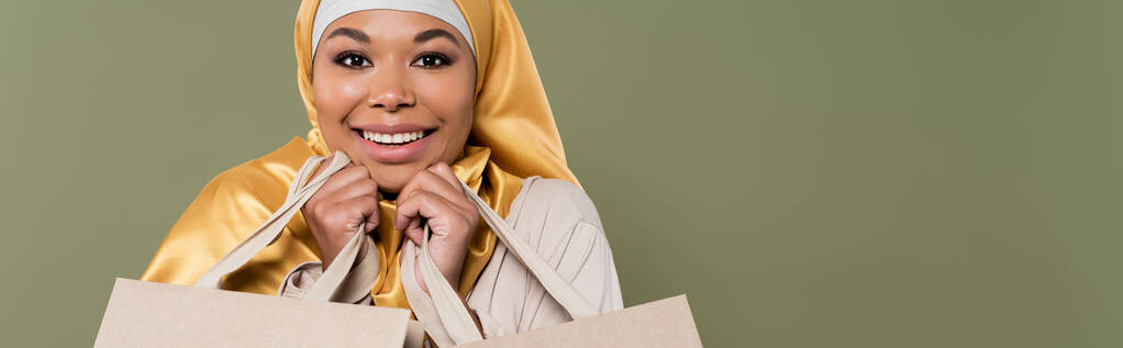 Mujer alegre multirracial en hijab dorado sosteniendo bolsas aisladas en verde, pancarta  - Foto, imagen