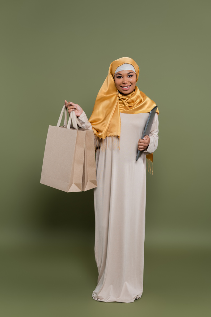 Comprimento total da mulher multicultural no hijab segurando laptop e sacos de compras no fundo verde - Foto, Imagem