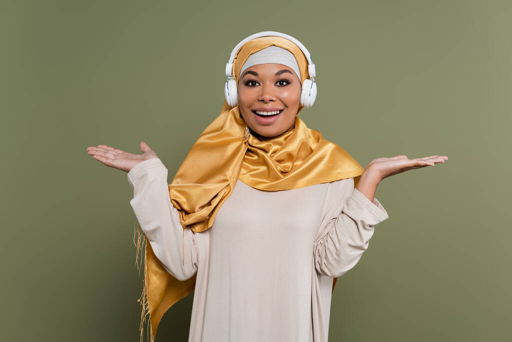 Θετική πολυφυλετική γυναίκα με μαντίλα και ακουστικά που δείχνουν με τα χέρια στο πράσινο φόντο - Φωτογραφία, εικόνα