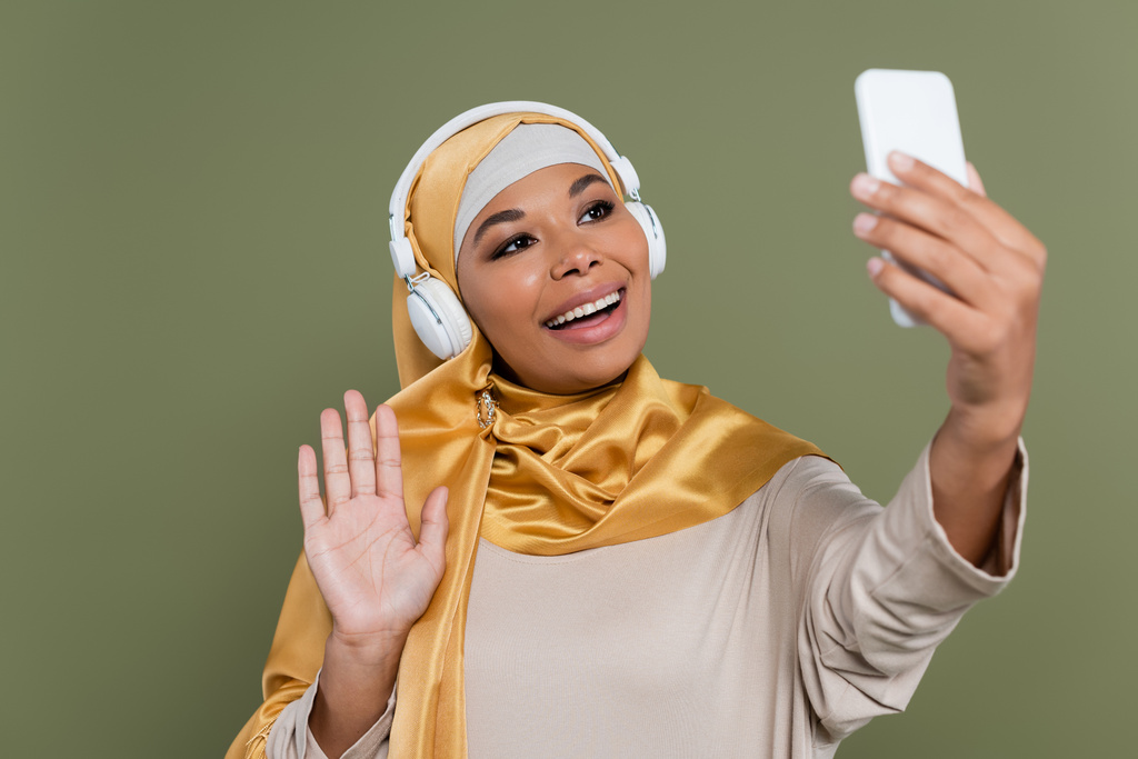 緑の上に隔離されたスマートフォンでビデオ通話を持つヒジャーブとヘッドフォンで陽気な多人種間の女性  - 写真・画像