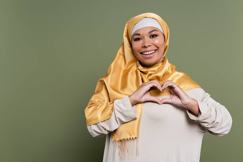 Mulher multirracial sorridente no hijab mostrando sinal de coração no fundo verde - Foto, Imagem