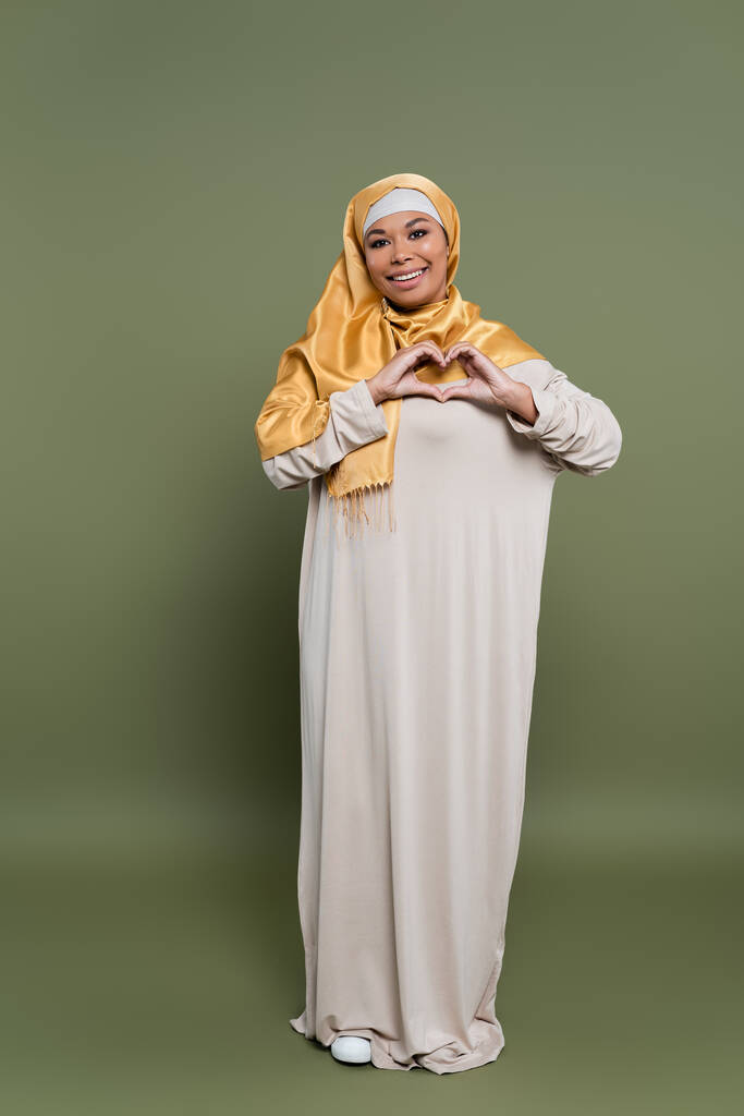 Carefree πολυφυλετική γυναίκα σε χιτζάμπ δείχνει σημάδι αγάπης με τα χέρια στο πράσινο φόντο - Φωτογραφία, εικόνα
