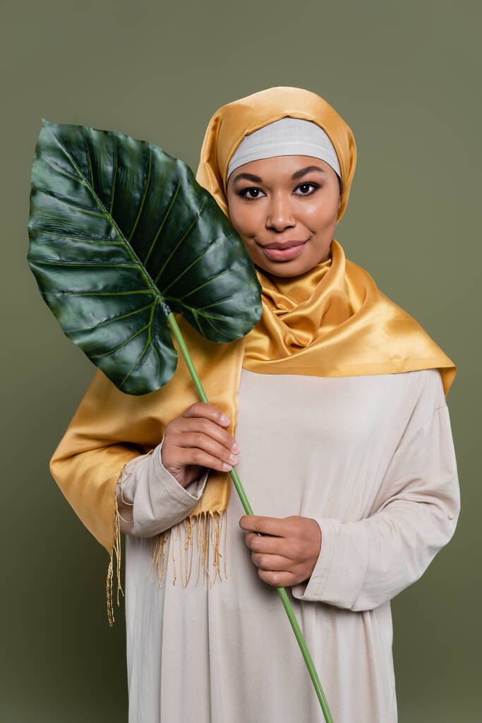 Νεαρή πολυφυλετική γυναίκα με μαντίλα κρατώντας τροπικό φύλλο σε πράσινο φόντο - Φωτογραφία, εικόνα
