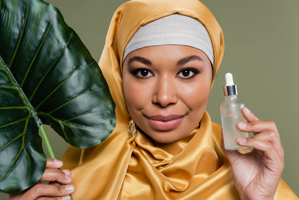 портрет мусульманської багаторасової жінки в жовтому шовковому хіджабі, що тримає косметичну сироватку і тропічний лист ізольовано на зеленому
 - Фото, зображення
