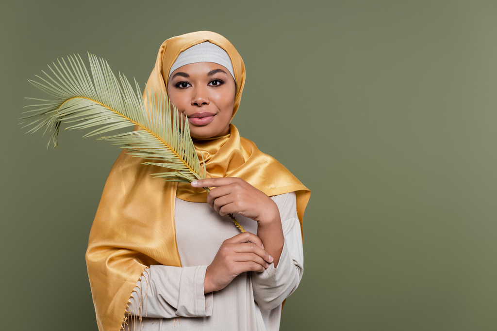 mujer multirracial feliz con maquillaje usando hiyab satinado amarillo y sosteniendo hoja exótica aislada en verde - Foto, Imagen