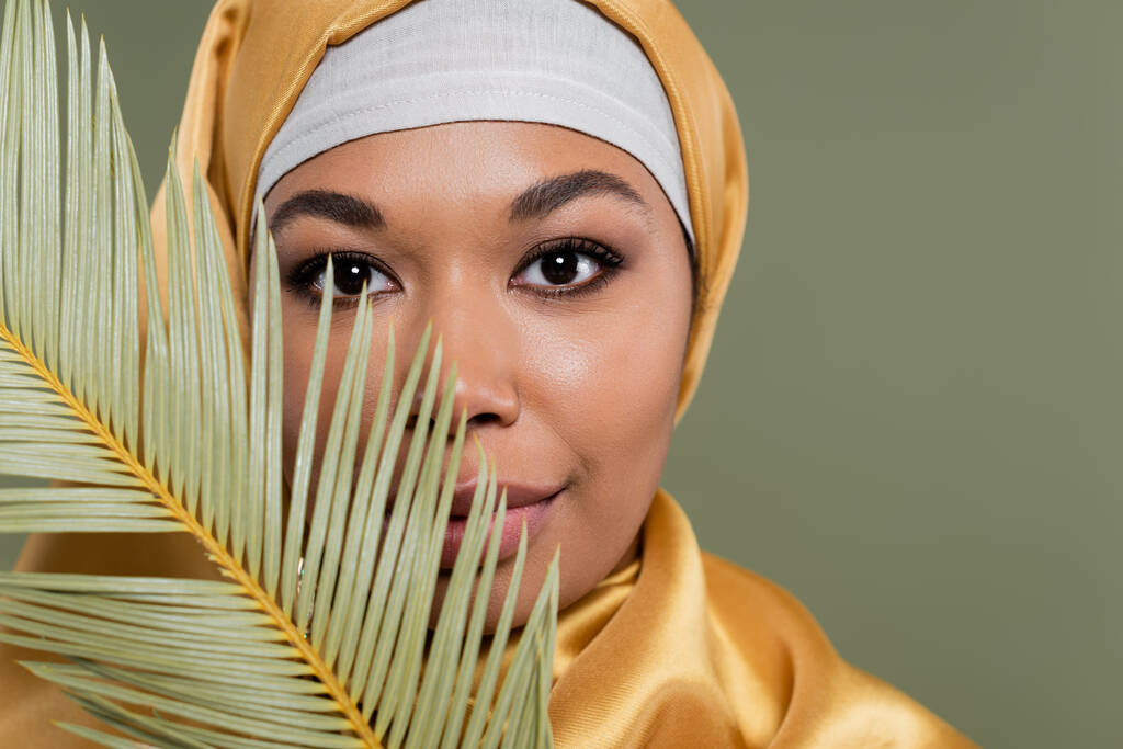 portrait of multiracial muslim woman with makeup looking at camera near tropical leaf isolated on green - Photo, Image