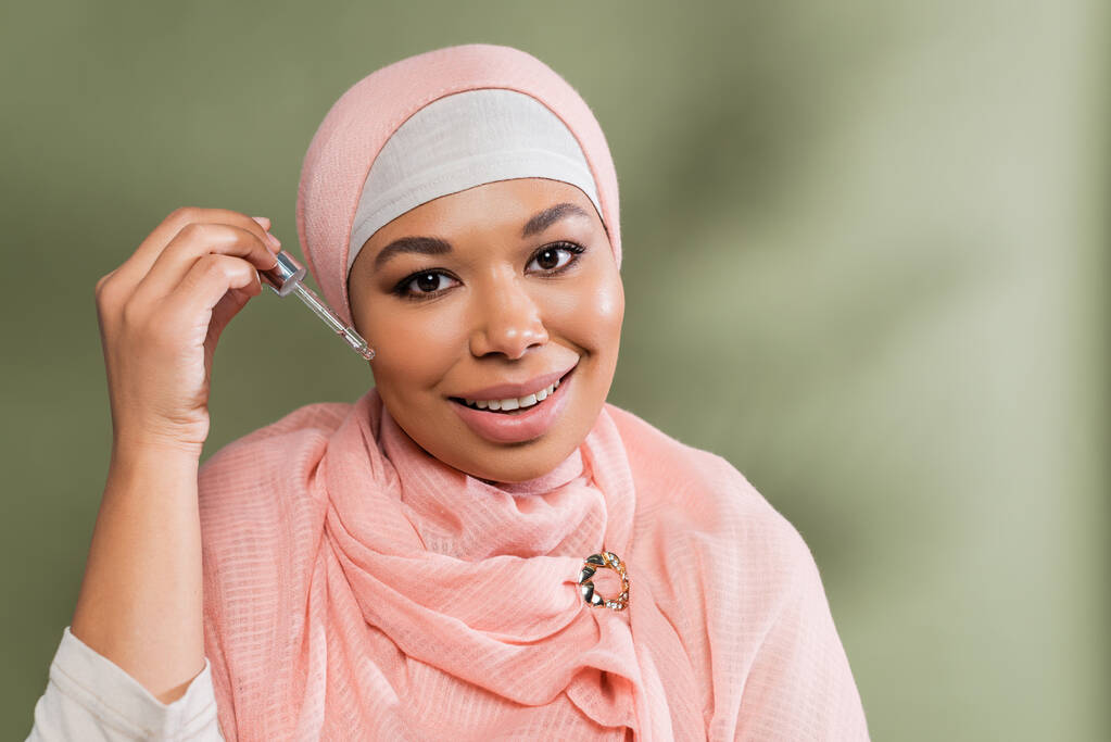 joyful multiracial muslim woman in pink hijab applying moisturizing serum on green background - Photo, Image