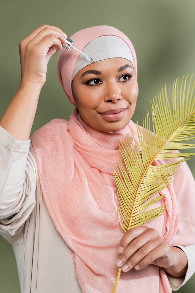 mulher muçulmana multirracial em hijab rosa segurando folha de planta exótica e aplicando soro hidratante em fundo verde - Foto, Imagem